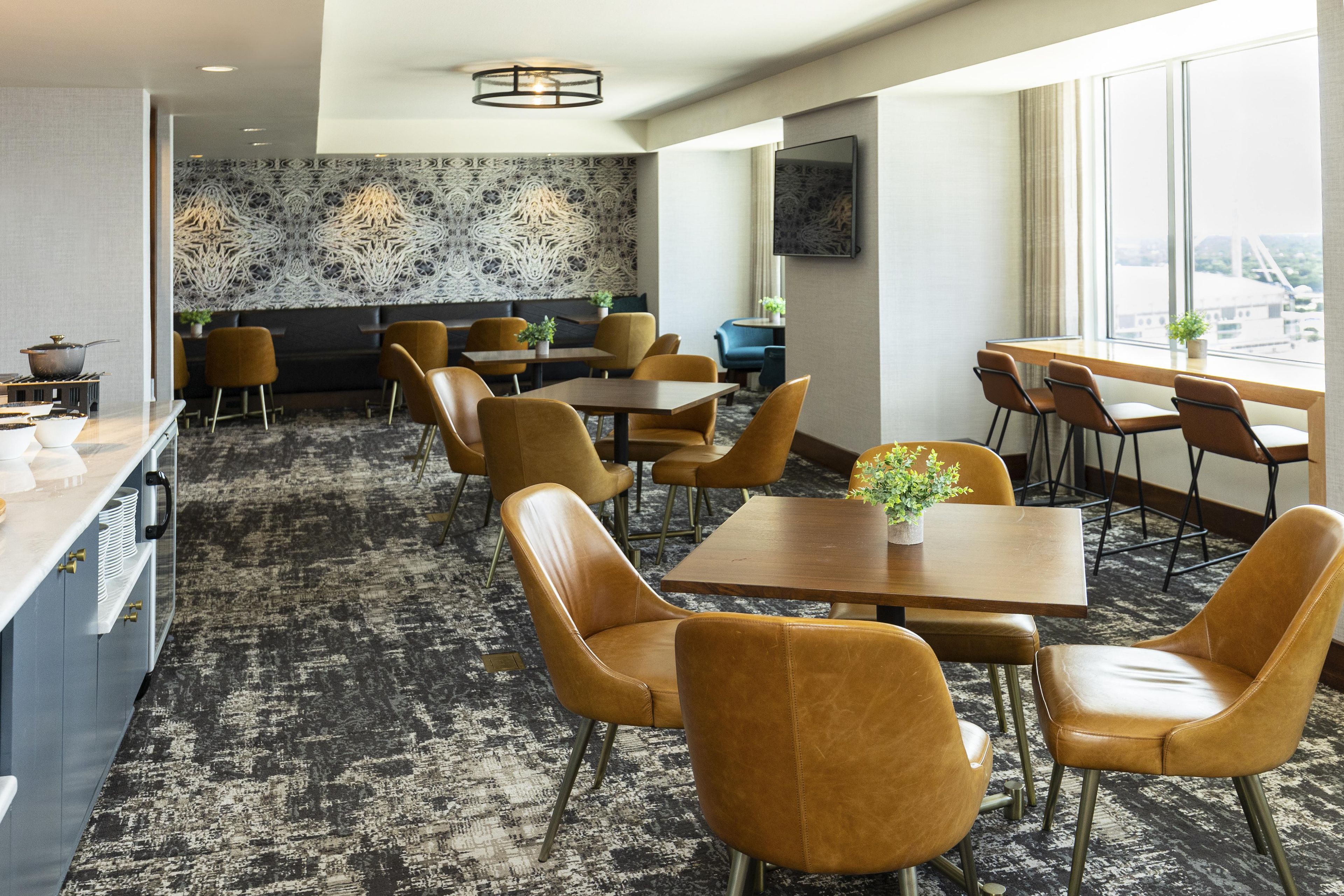 A hotel dining area with multiple tables and chairs, a patterned wall, and a view through large windows.