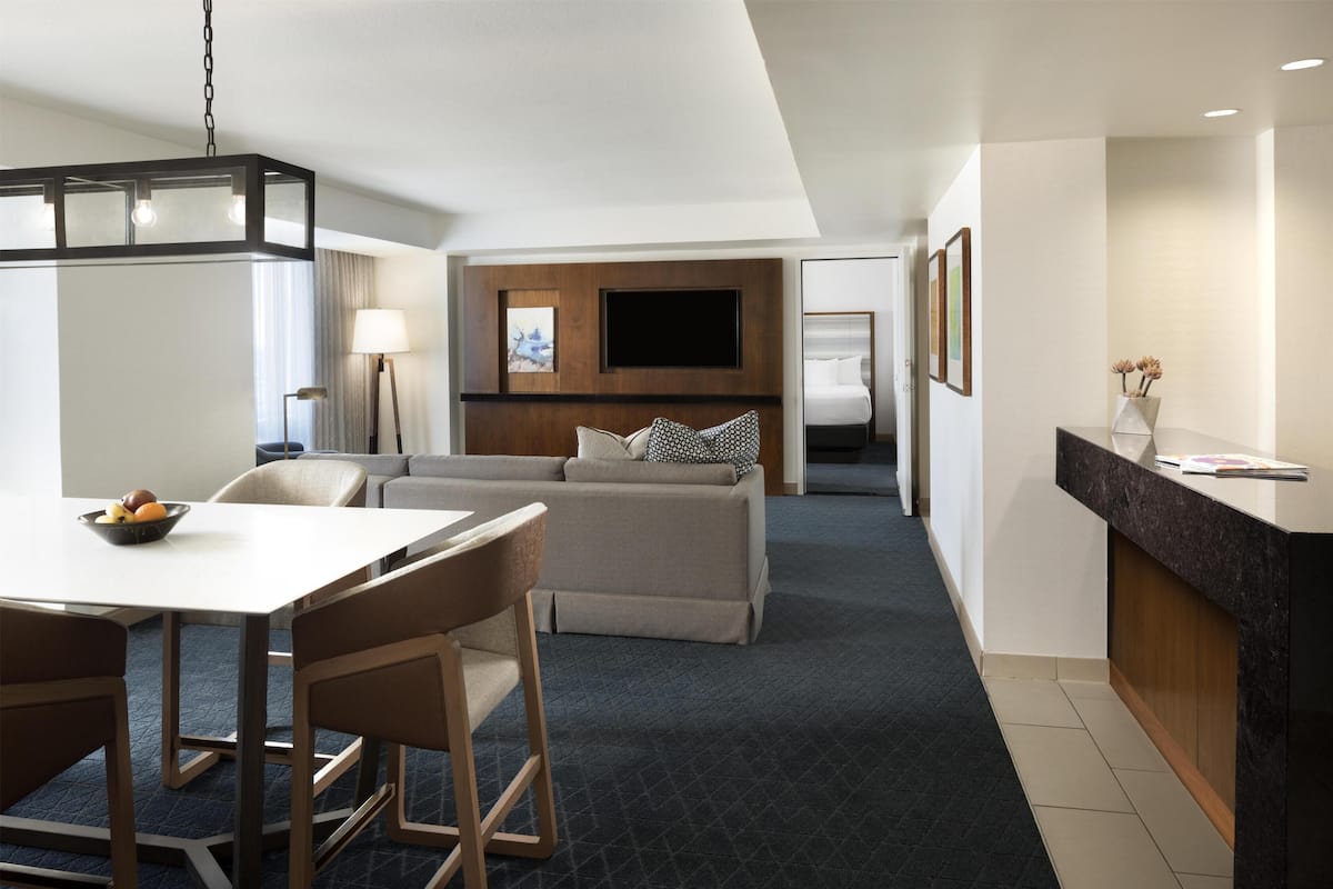 A modern hotel room with a dining area, a sofa, a flat-screen TV, and a small kitchen counter.