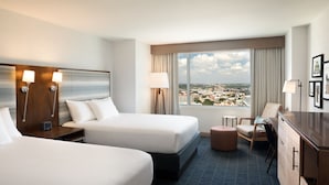 Habitación de hotel con dos camas, un escritorio, una silla, vista a la ciudad y un ventanal con cortinas.