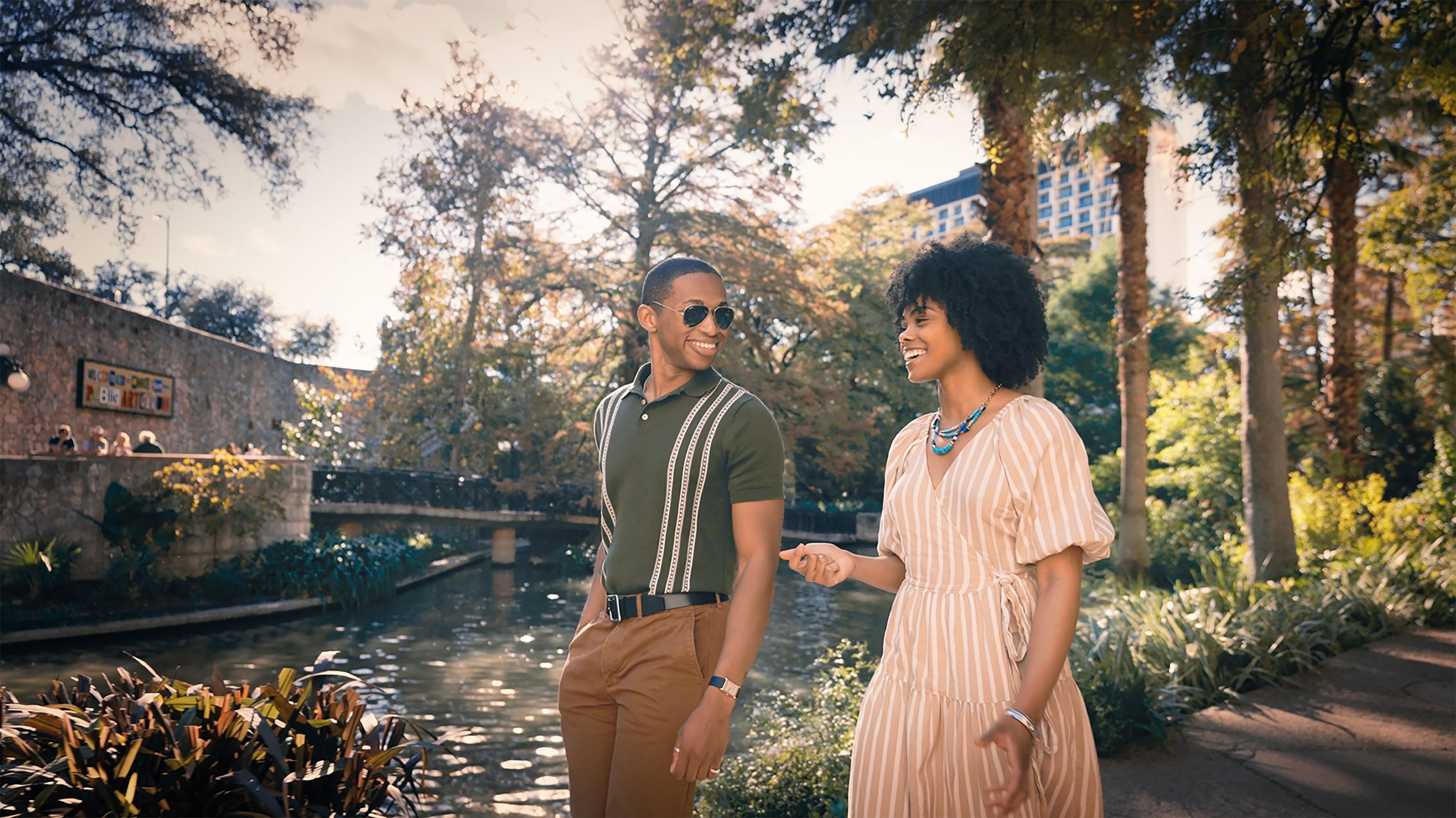 A couple standing by a canal with trees and a stone wall in the background.