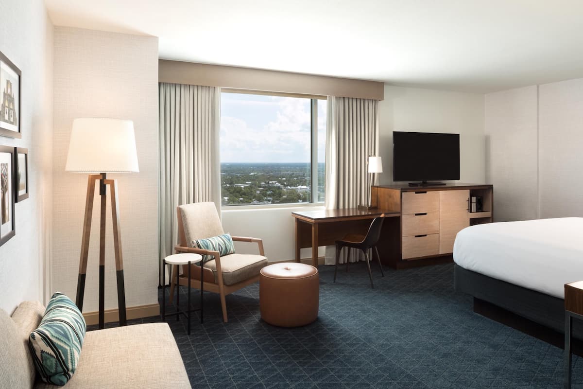 A hotel room with a bed, a desk with a chair, a television, a lamp, and a view of the city.