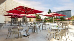 An outdoor patio area with multiple round tables and chairs, red umbrellas, and a stone building in the background. - Grand Hyatt San Antonio River Walk (San Antonio)