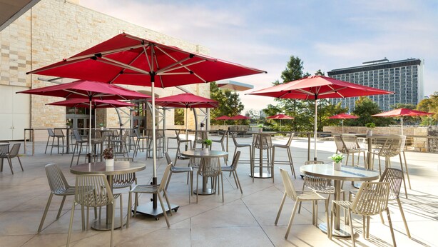 An outdoor patio area with multiple round tables and chairs, red umbrellas, and a stone building in the background. - Grand Hyatt San Antonio River Walk (San Antonio)
