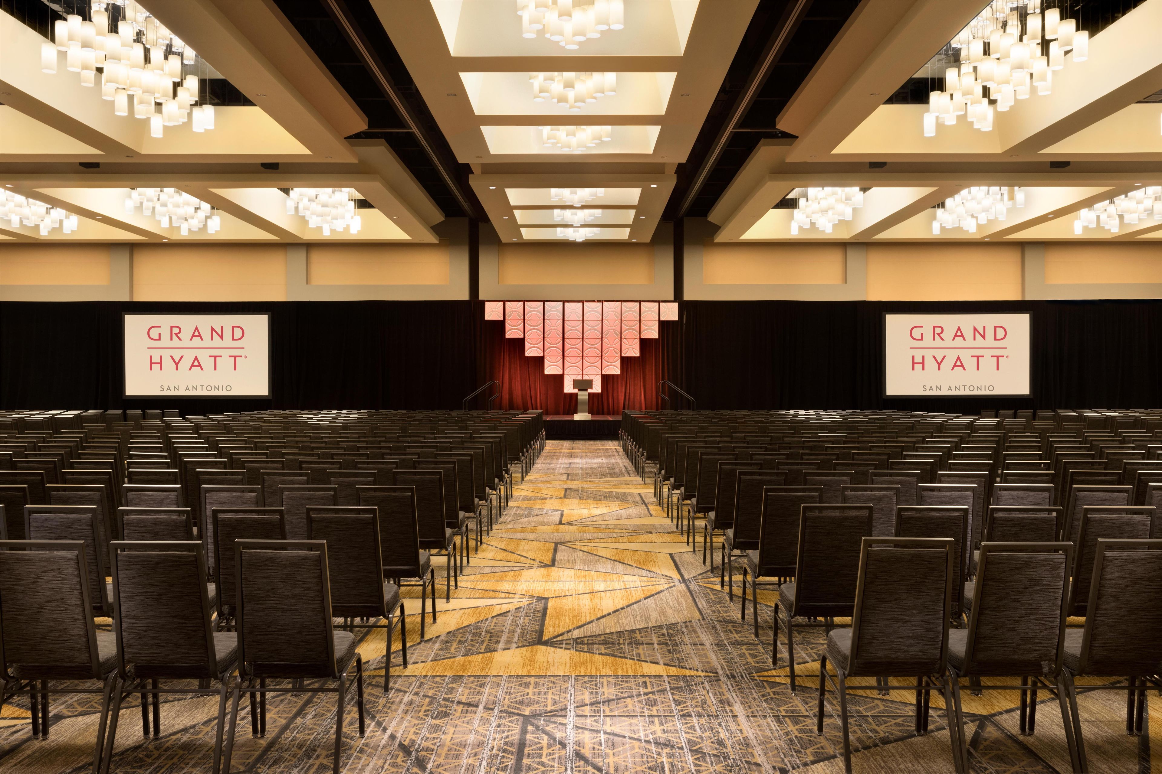 Un salón de conferencias con filas de sillas, una alfombra estampada y un escenario con cortina roja.
