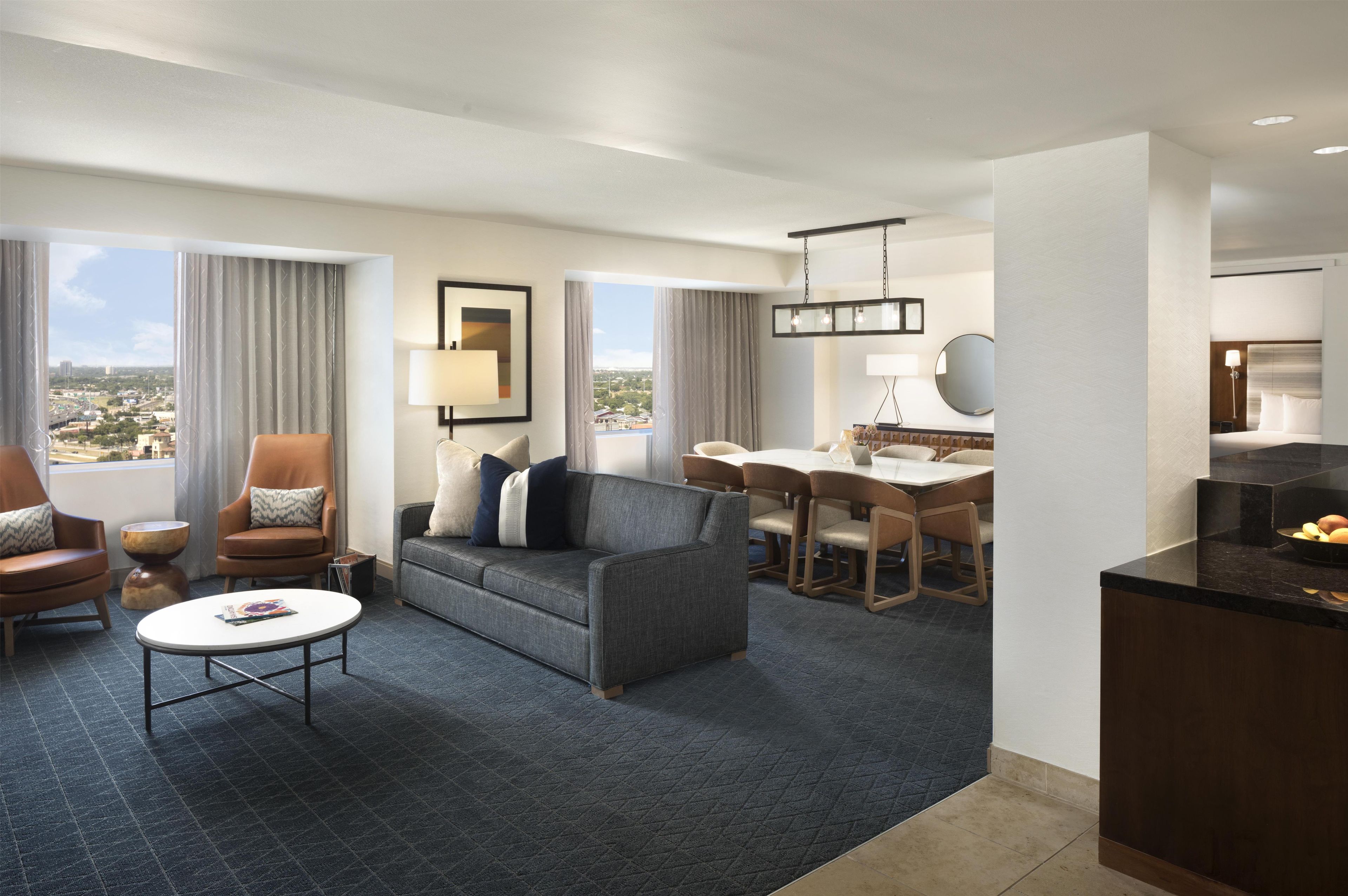 Habitación de hotel con sofá, sillas, una mesa de comedor, zona de cocina y ventanas amplias con vista a la ciudad.