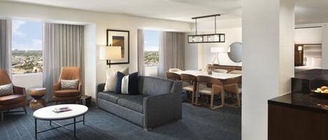 Habitación de hotel con sofá, sillas, una mesa de comedor, zona de cocina y ventanas amplias con vista a la ciudad.