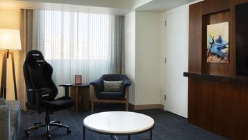 A hotel room with a black gaming chair, a round table, a wooden reception desk, a painting on the wall, and a window with sheer curtains.