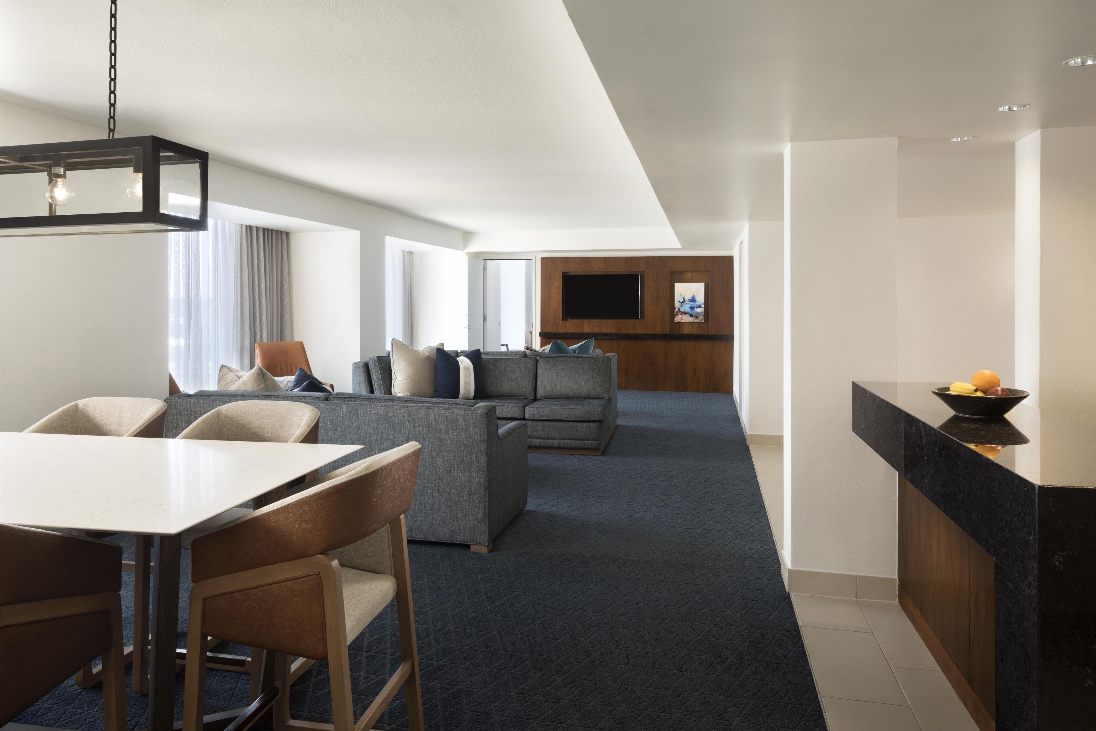 A modern hotel room with a dining area, a sofa set, a flat-screen TV, and a kitchen counter.