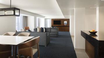 A modern hotel room with a dining area, a sofa set, a flat-screen TV, and a kitchen counter.