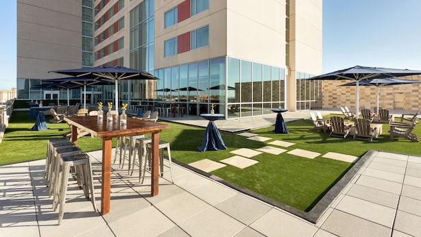 Un patio al aire libre con mesas, sillas y sombrillas, rodeado por un edificio con grandes ventanales y un césped.
