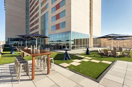 An outdoor patio area with tables, chairs, and umbrellas, surrounded by a building with large windows and a grassy lawn.