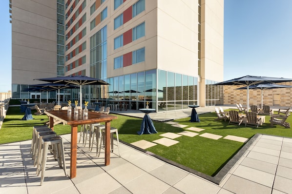 Un patio al aire libre con mesas, sillas y sombrillas, rodeado por un edificio con grandes ventanales y un césped.