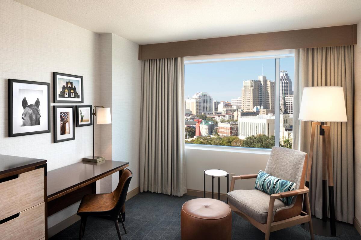 A hotel room with a desk, chair, ottoman, and a view of the cityscape through the window.