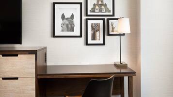 Habitación de hotel con un escritorio de madera, una silla negra, un reposapiés marrón y cuadros en la pared.