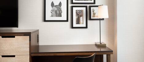 Habitación de hotel con un escritorio de madera, una silla negra, un reposapiés marrón y cuadros en la pared.