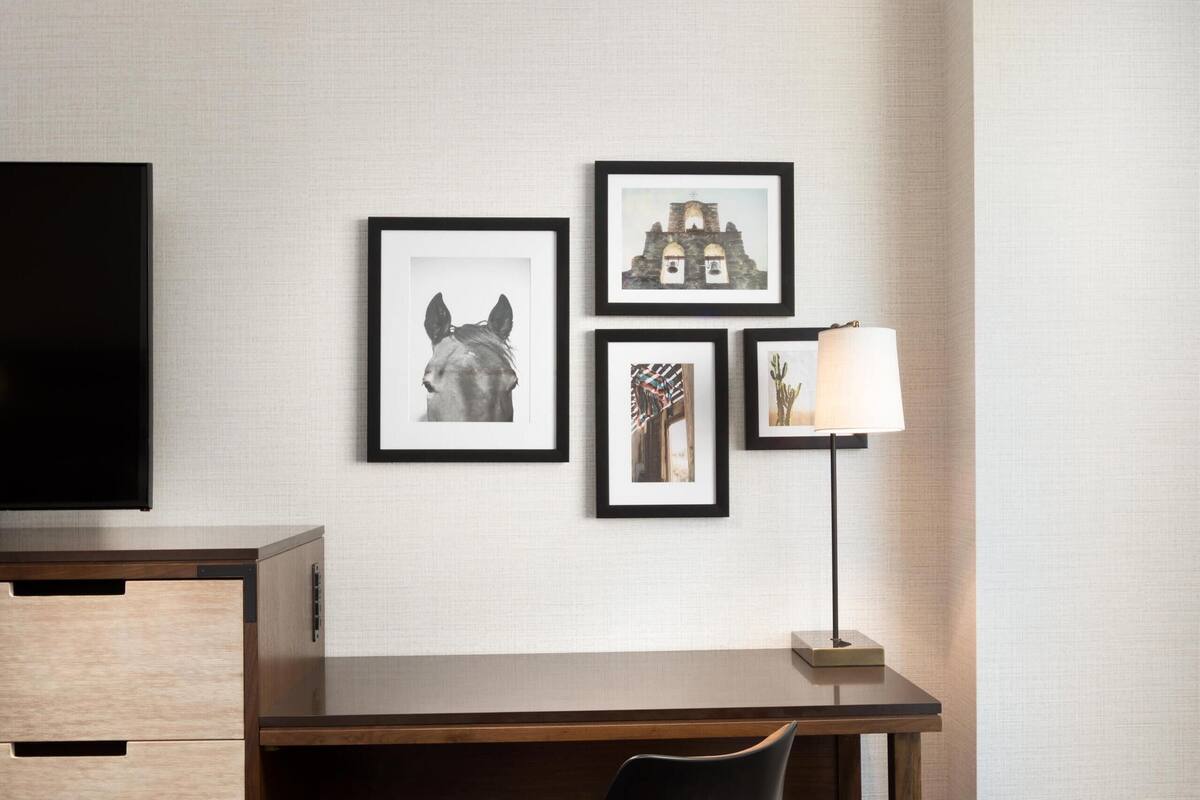 A hotel room with a wooden desk, a black chair, a brown ottoman, and framed artwork on the wall.
