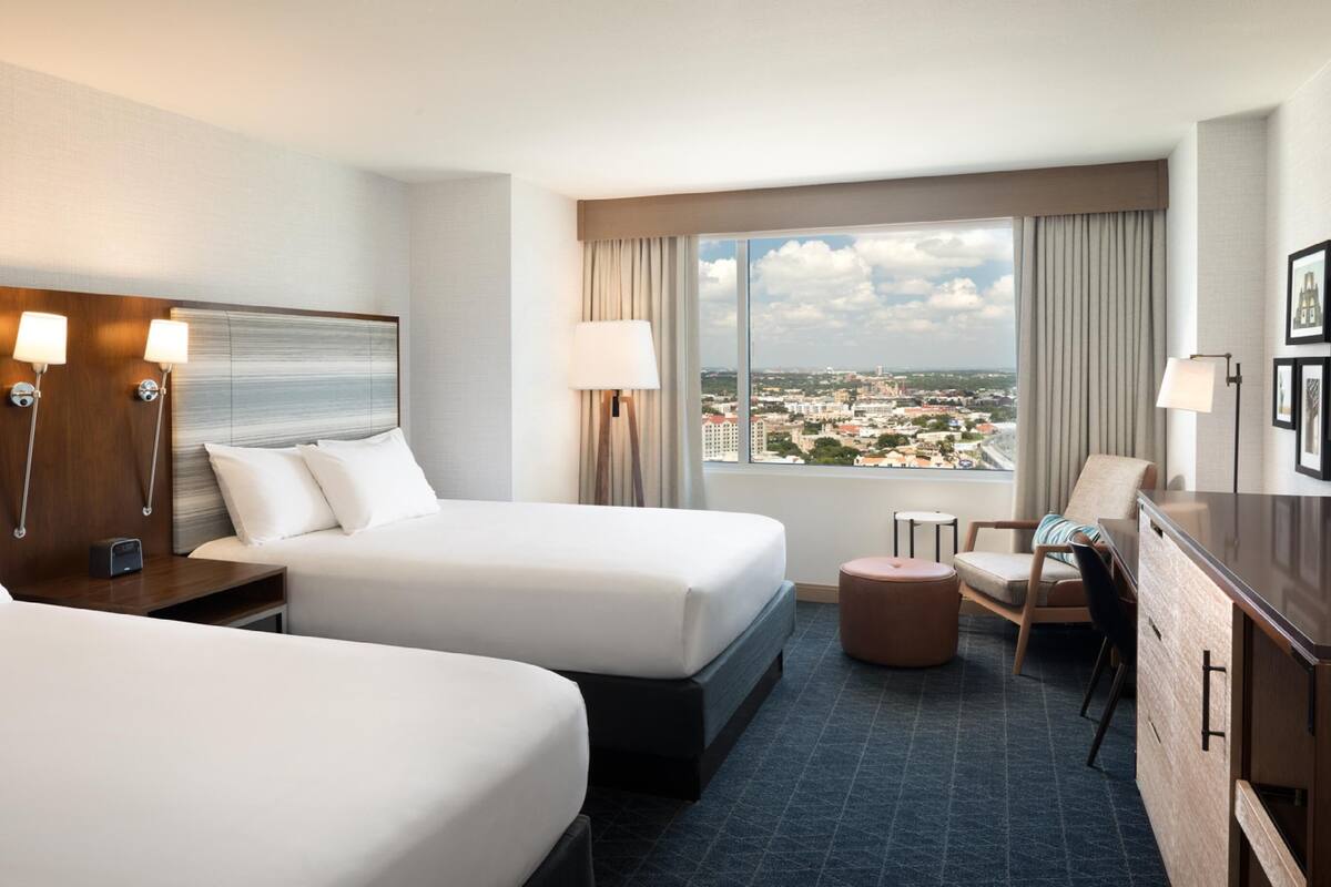 A hotel room with two beds, a desk, a chair, a small table, and a view of a cityscape.