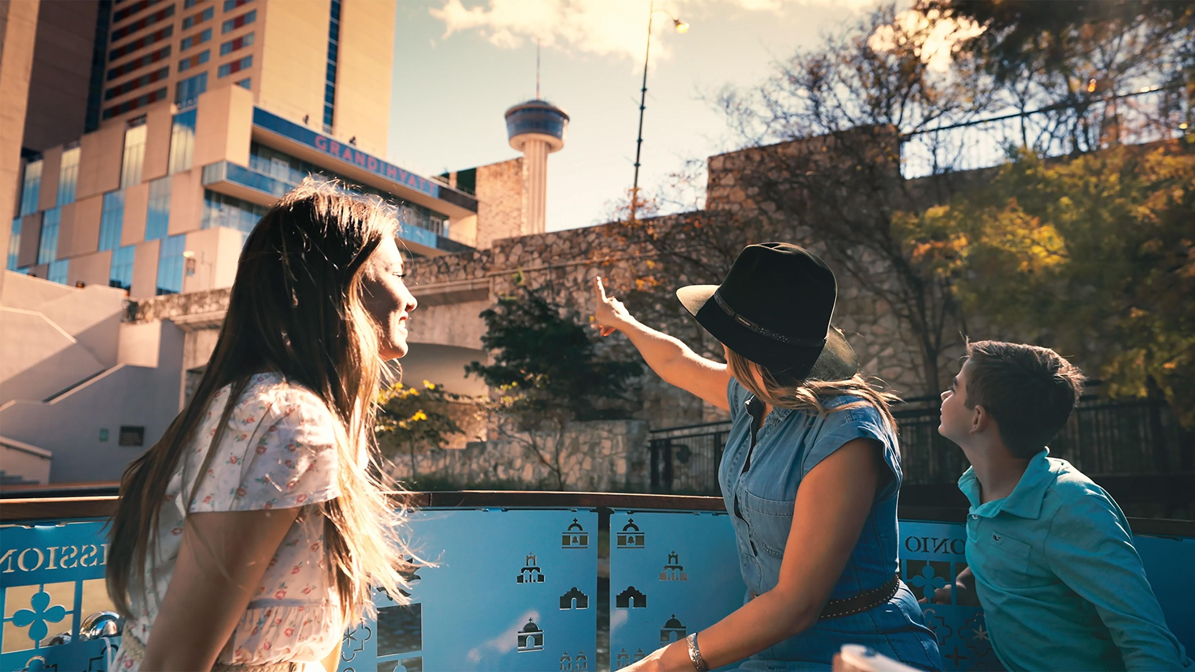 A group of people standing near a blue barrier with icons, a tall building with a tower in the background, and a clear sky.