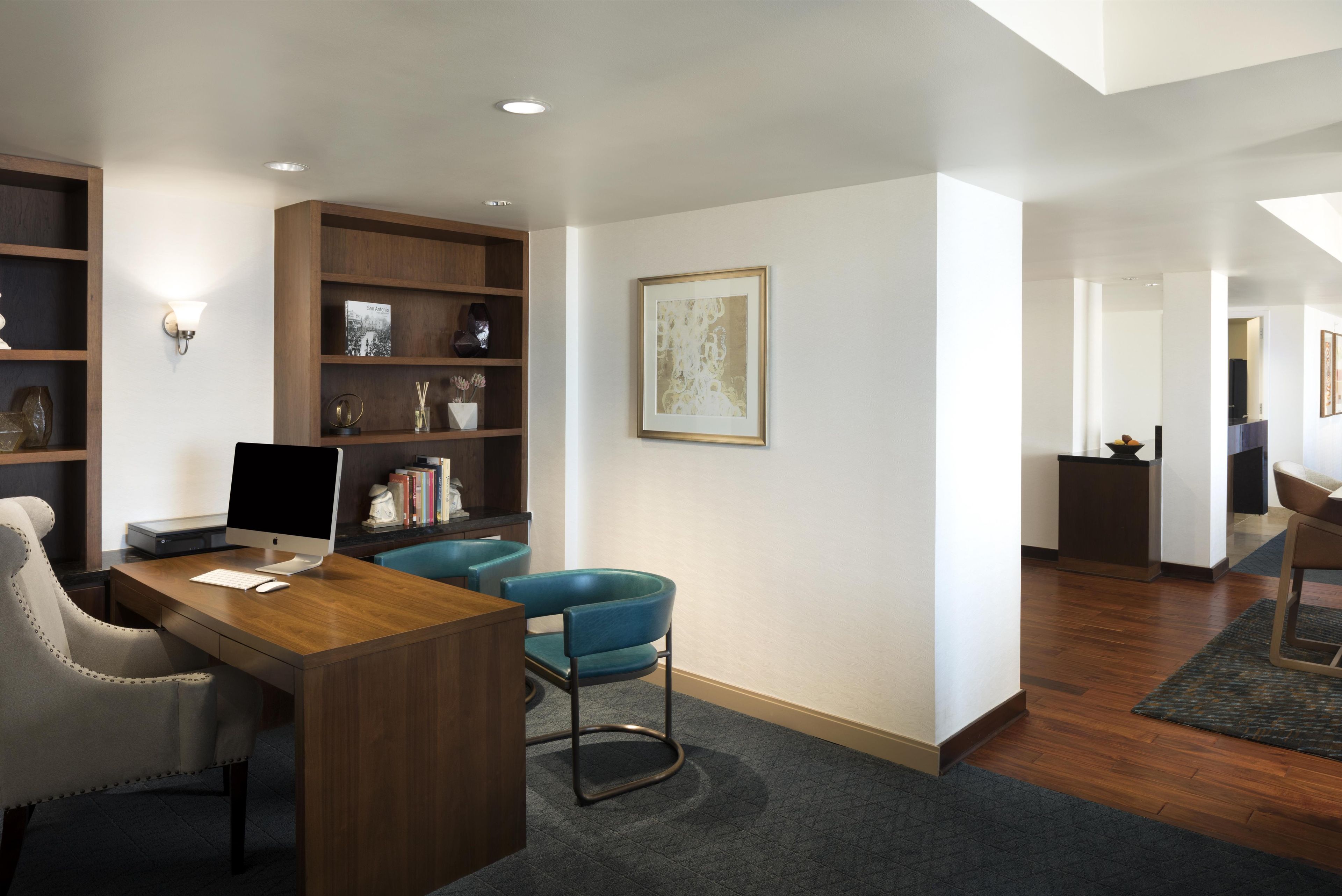 A modern office space with a desk, computer, chairs, shelves with decorative items, and a framed artwork on the wall.