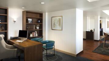 A modern office space with a desk, computer, chairs, shelves with decorative items, and a framed artwork on the wall.