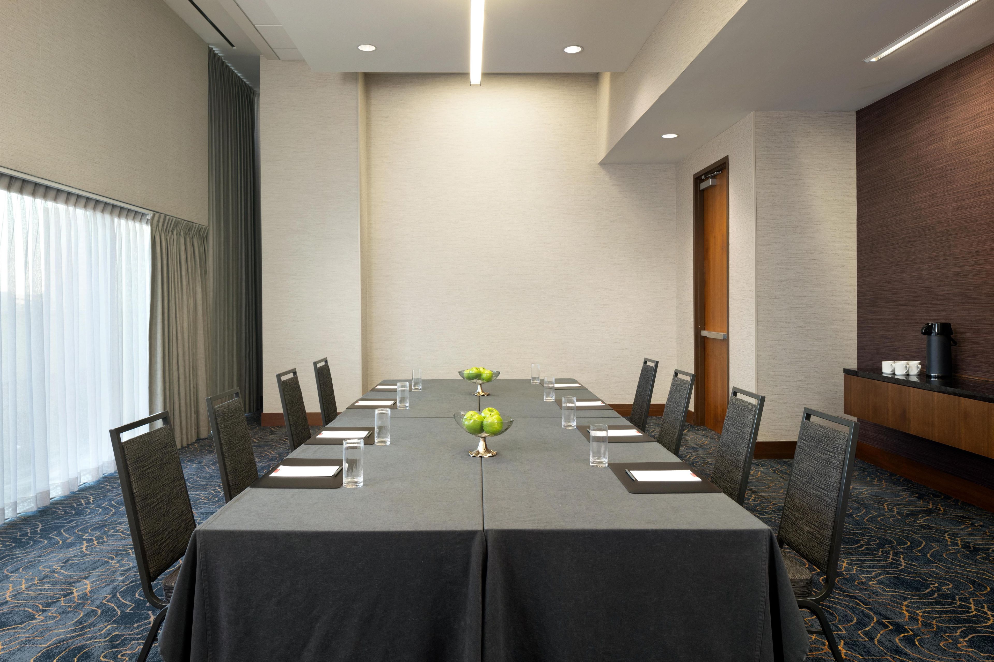 A conference room with a long table, chairs, a coffee station, and a window with curtains.