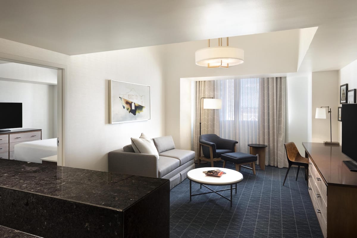 A modern hotel room with a kitchen area, a sofa, a dining table, and a TV.