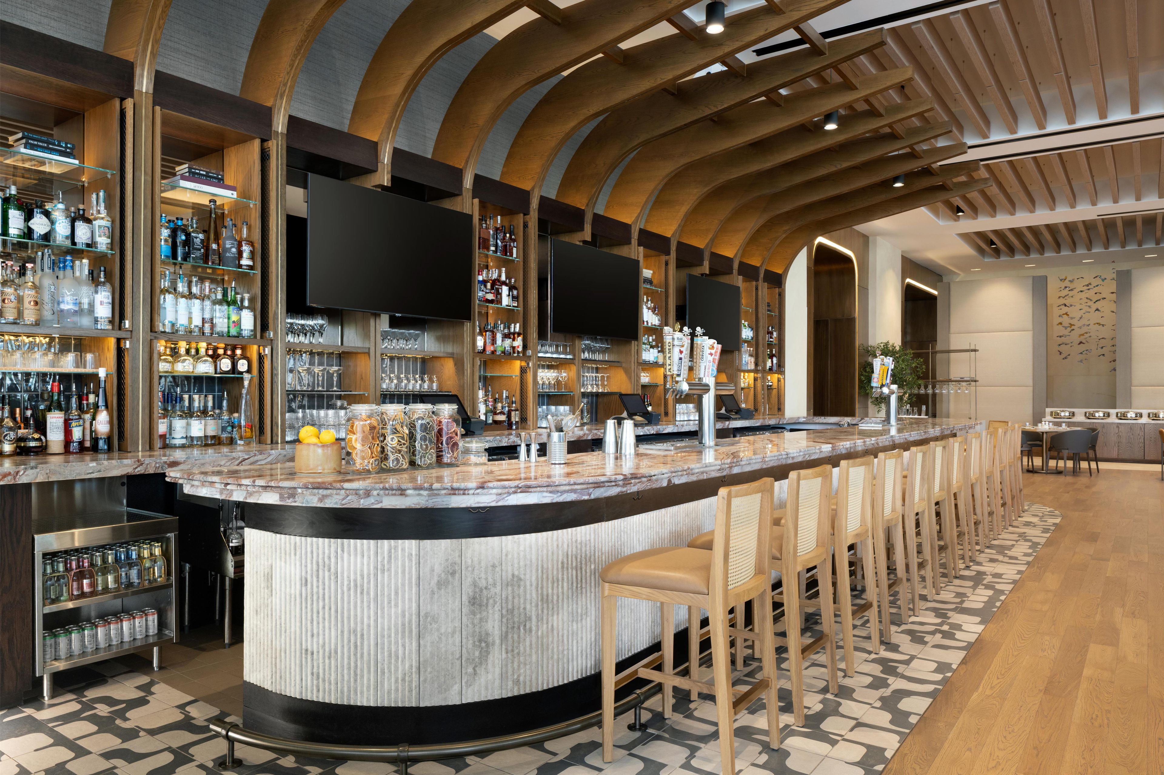 A bar area with a curved counter, bar stools, shelves with bottles, glasses, and a flat-screen TV mounted on the wall.