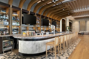 A bar area with a curved counter, bar stools, shelves with bottles, glasses, and a flat-screen TV mounted on the wall. - Grand Hyatt San Antonio River Walk (San Antonio)