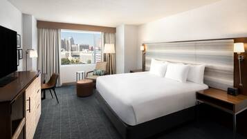 A hotel room with a large bed, a desk, a chair, a TV, and a view of the city.