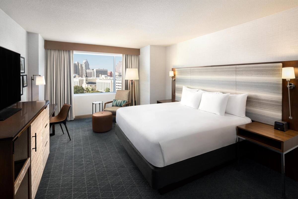 A hotel room with a large bed, a desk, a chair, a TV, and a view of the city.