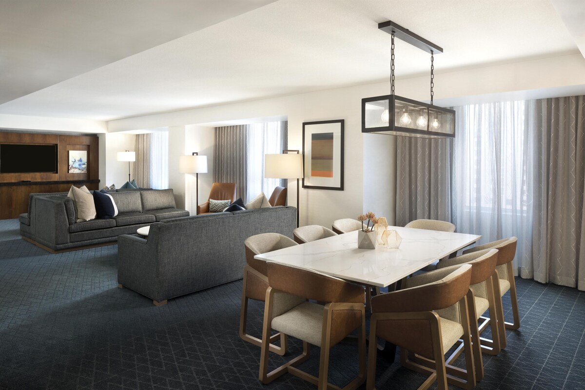 A modern hotel room with a dining area, sofa, and a flat-screen TV.