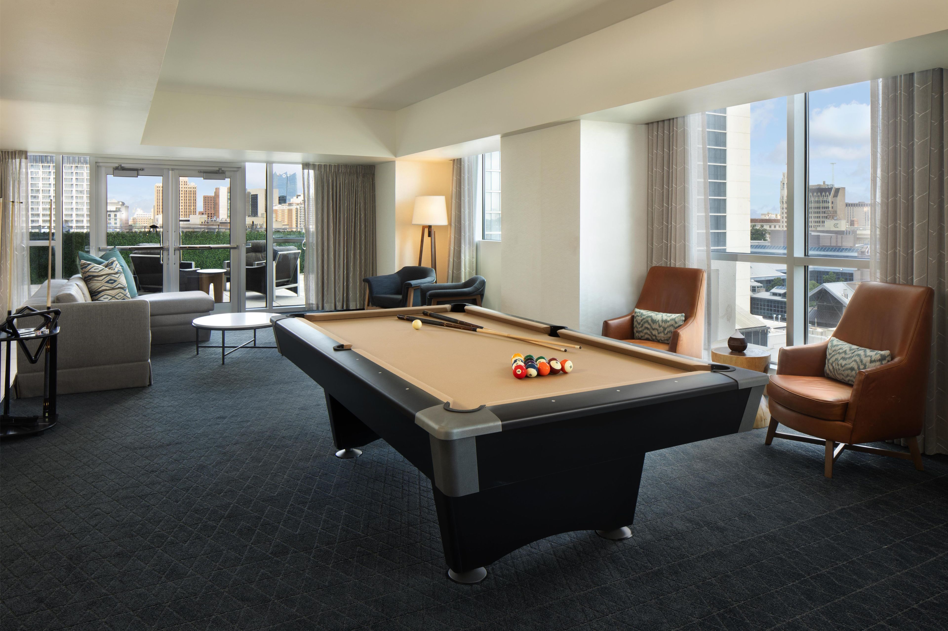 Una habitación de hotel con mesa de billar, sofá, dos sillones y vista a la ciudad.