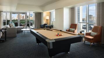 Una habitación de hotel con mesa de billar, sofá, dos sillones y vista a la ciudad.