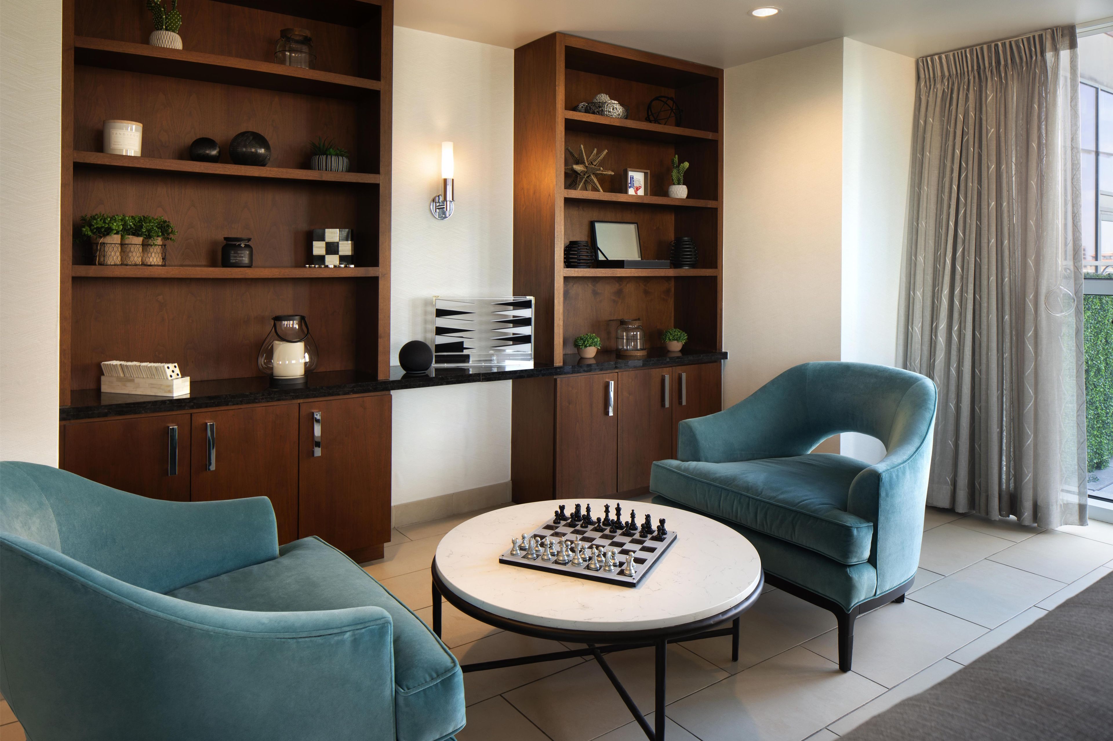 A living room with a chessboard, two teal armchairs, wooden shelving with various items, and a large window with curtains.