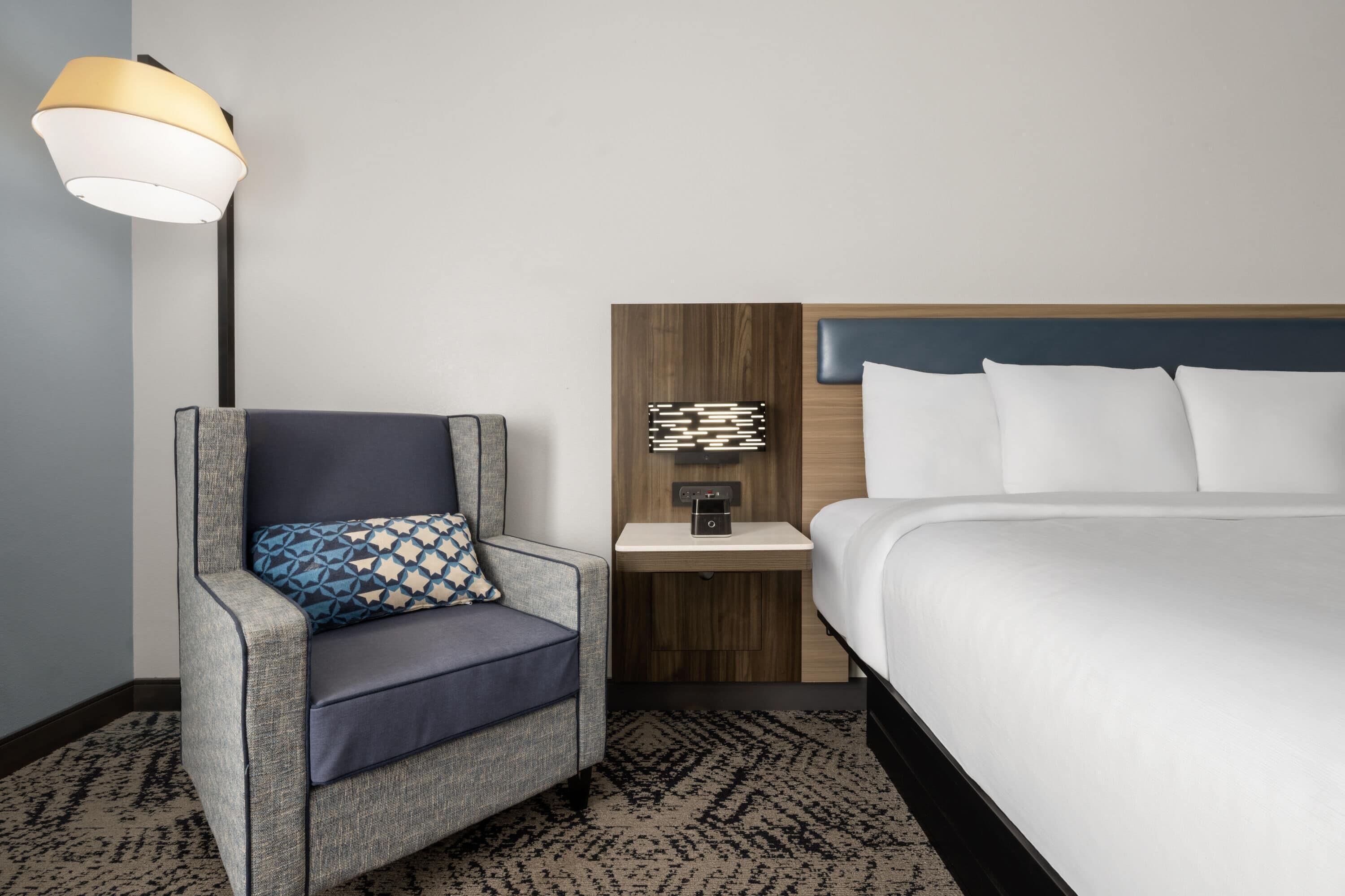 A hotel room with a bed, a chair with a patterned cushion, a nightstand with a lamp, and a wall-mounted artwork.