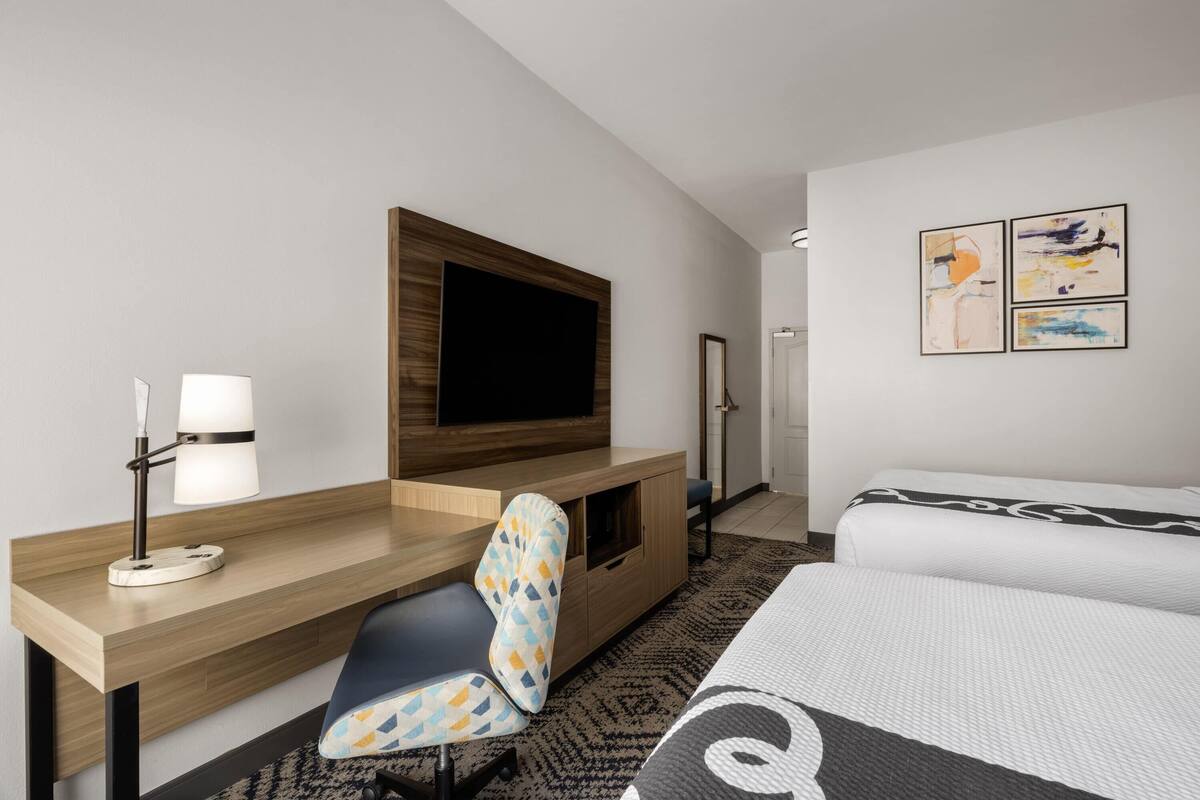 Una habitación de hotel con un escritorio de madera, un televisor, una cama, una silla y obras de arte en la pared.