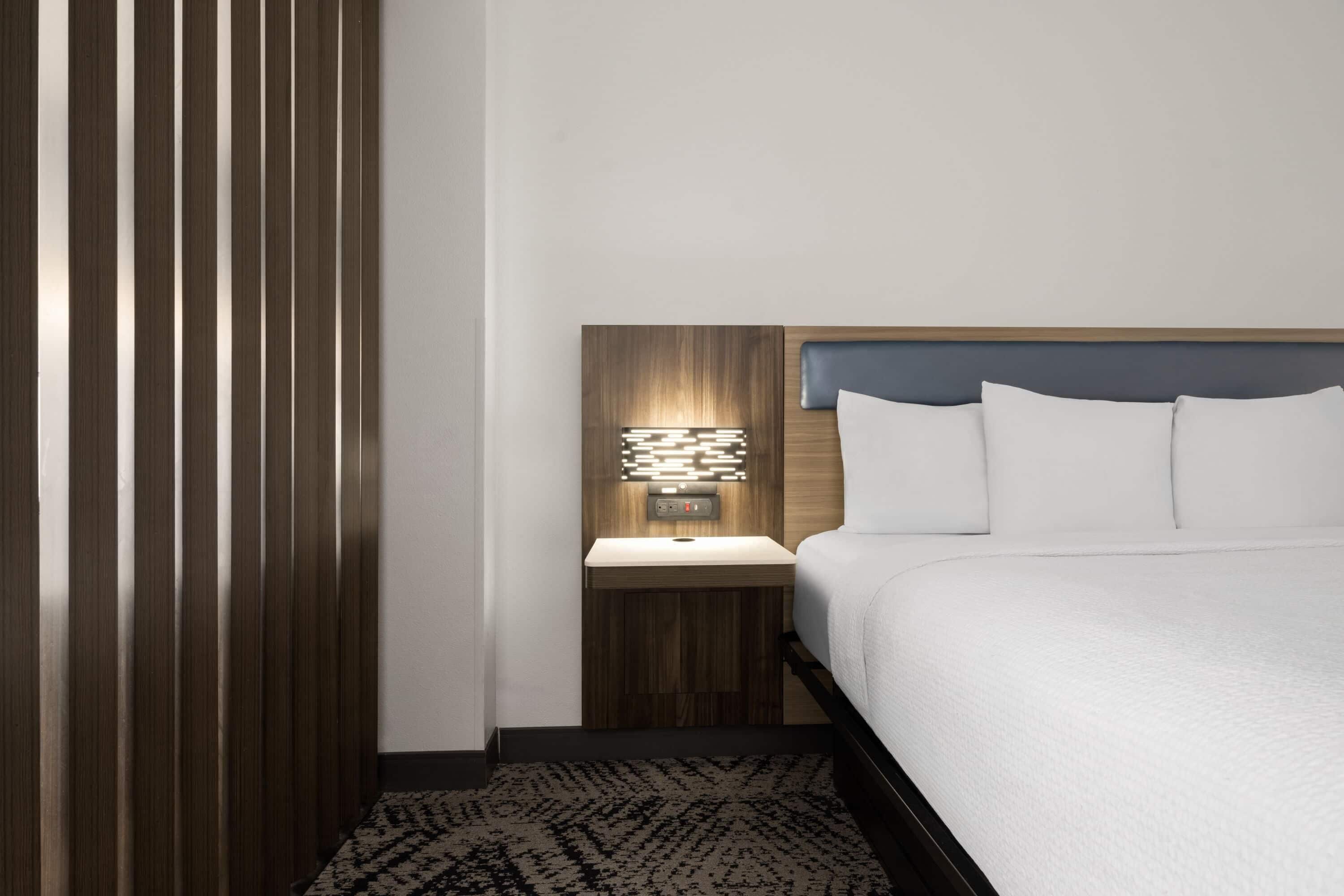 Habitación de hotel con cama, mesita de noche con lámpara, pared de paneles de madera y alfombra estampada.
