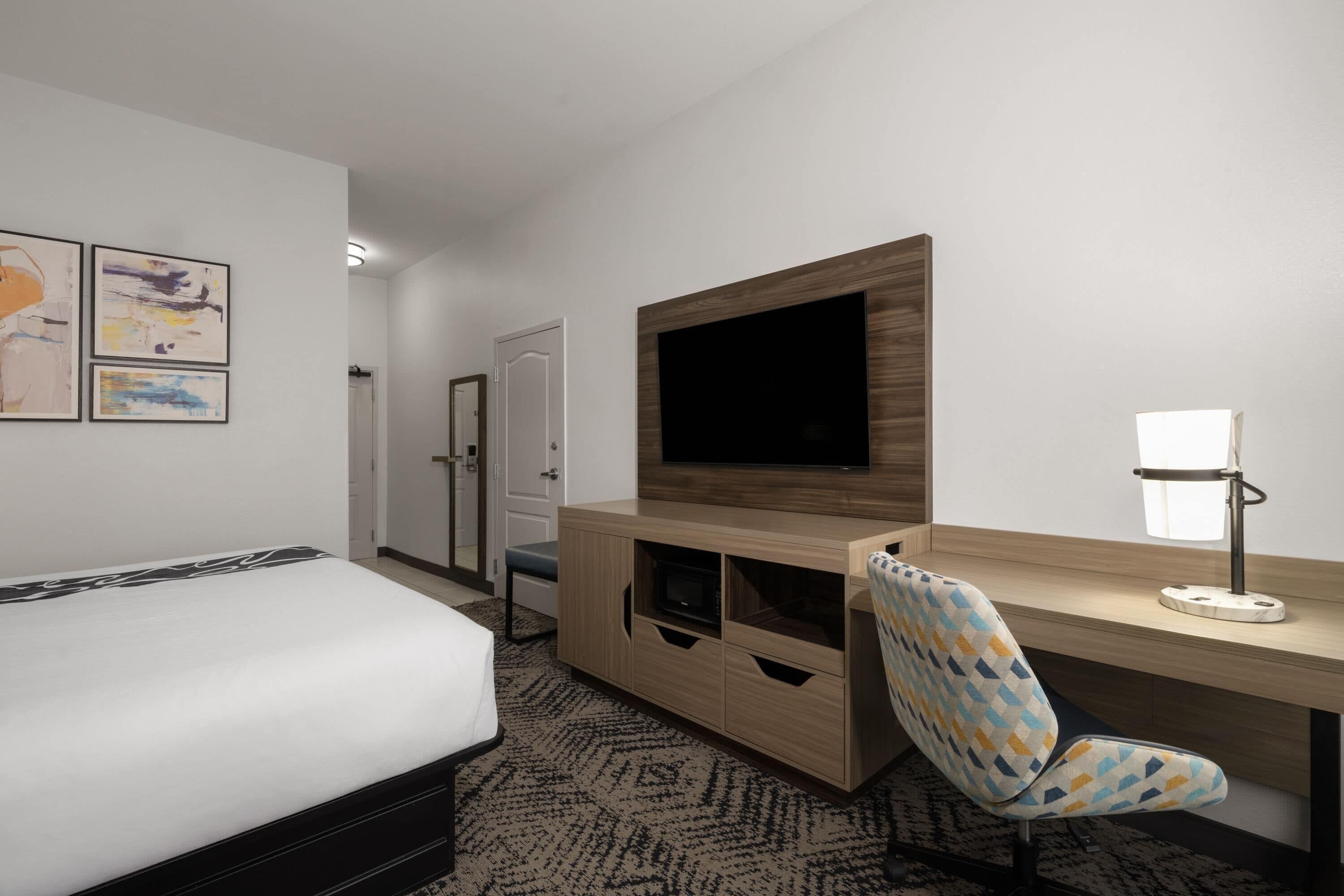 A hotel room with a bed, a TV mounted on a wooden cabinet, a desk with a lamp, and a patterned carpet.