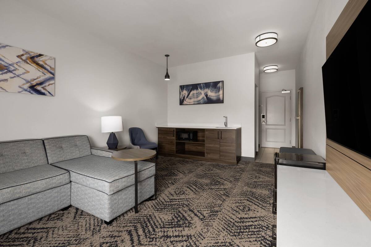 Una sala moderna con un sofá seccional gris, una mesita redonda pequeña, un sillón azul, un mueble con microondas y un televisor.