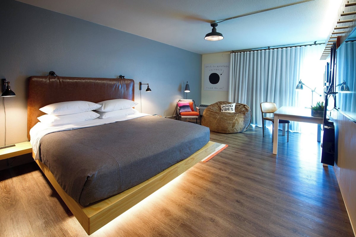 A modern hotel room with a large bed, a desk, a chair, and a wall-mounted TV.
