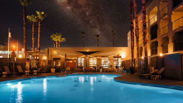Una piscina con sillones de playa y palmeras, rodeada por un edificio con balcones y luces colgantes.