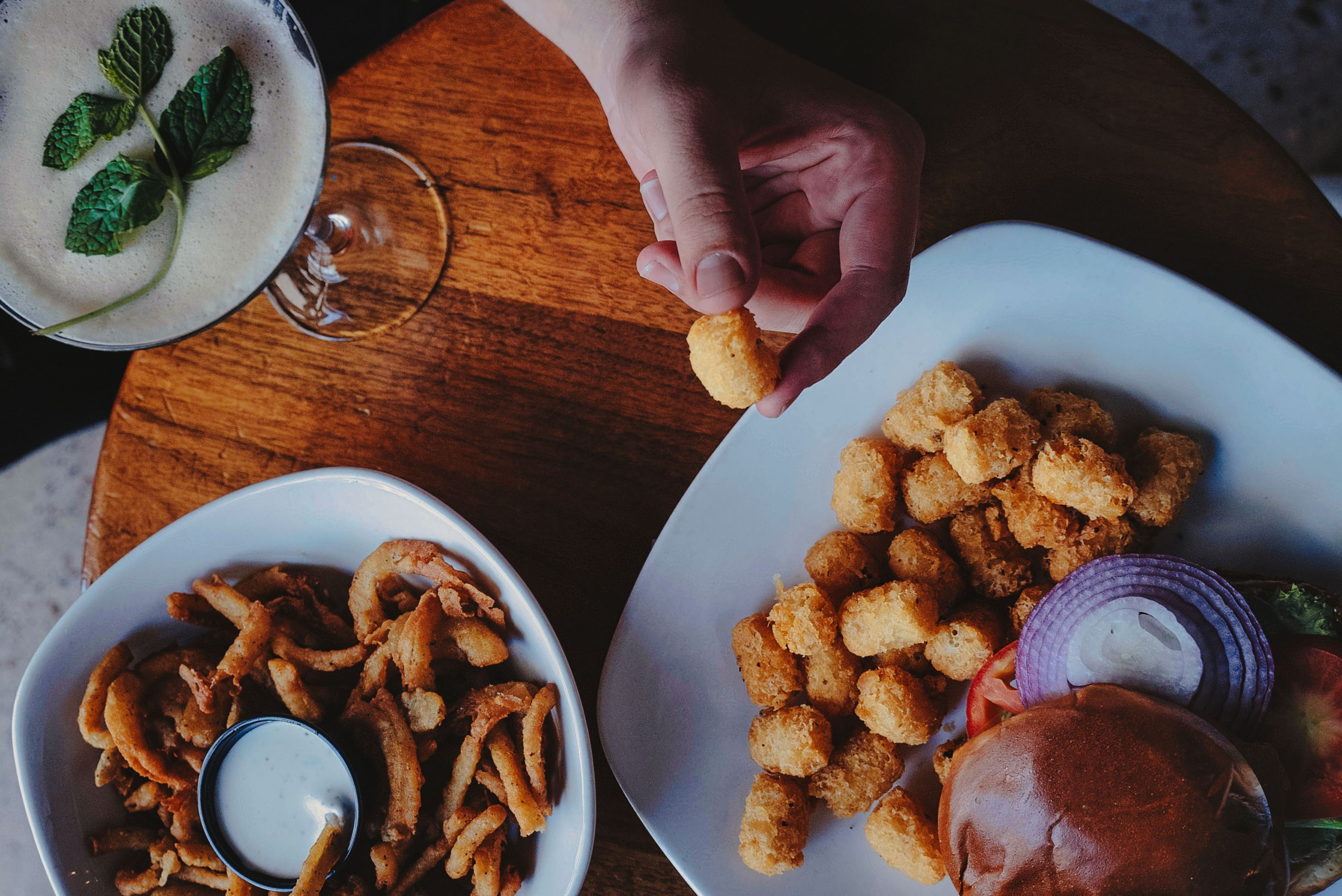 A table with a plate of tater tots, onion rings, and a burger, a glass of a creamy beverage, and a hand reaching for a tater tot.
