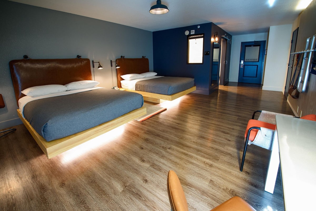 A hotel room with two beds, a desk, and a chair. There is a window providing natural light, and the room has wooden flooring.
