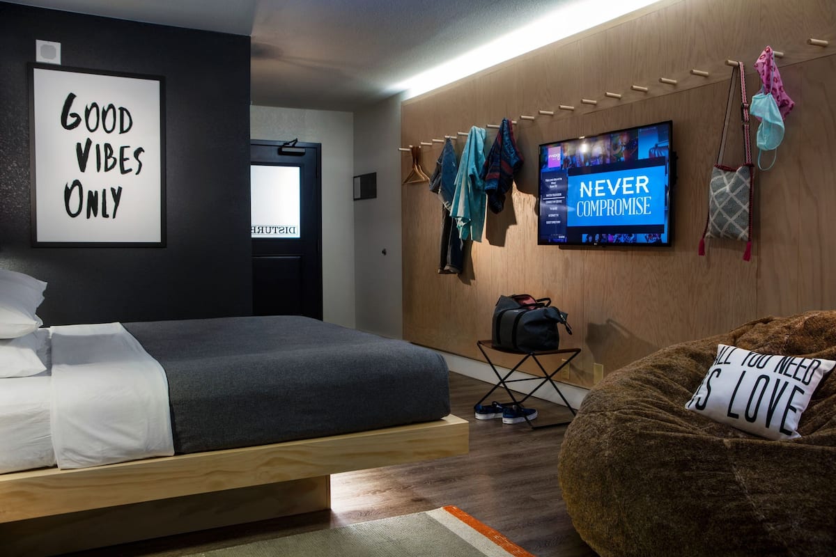 A modern hotel room with a bed, a bean bag chair, a TV, and motivational wall art.