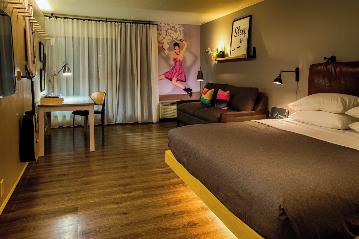 A hotel room with a wooden floor, a bed with a brown headboard, a desk with a chair, a couch with cushions, and a wall with a poster.