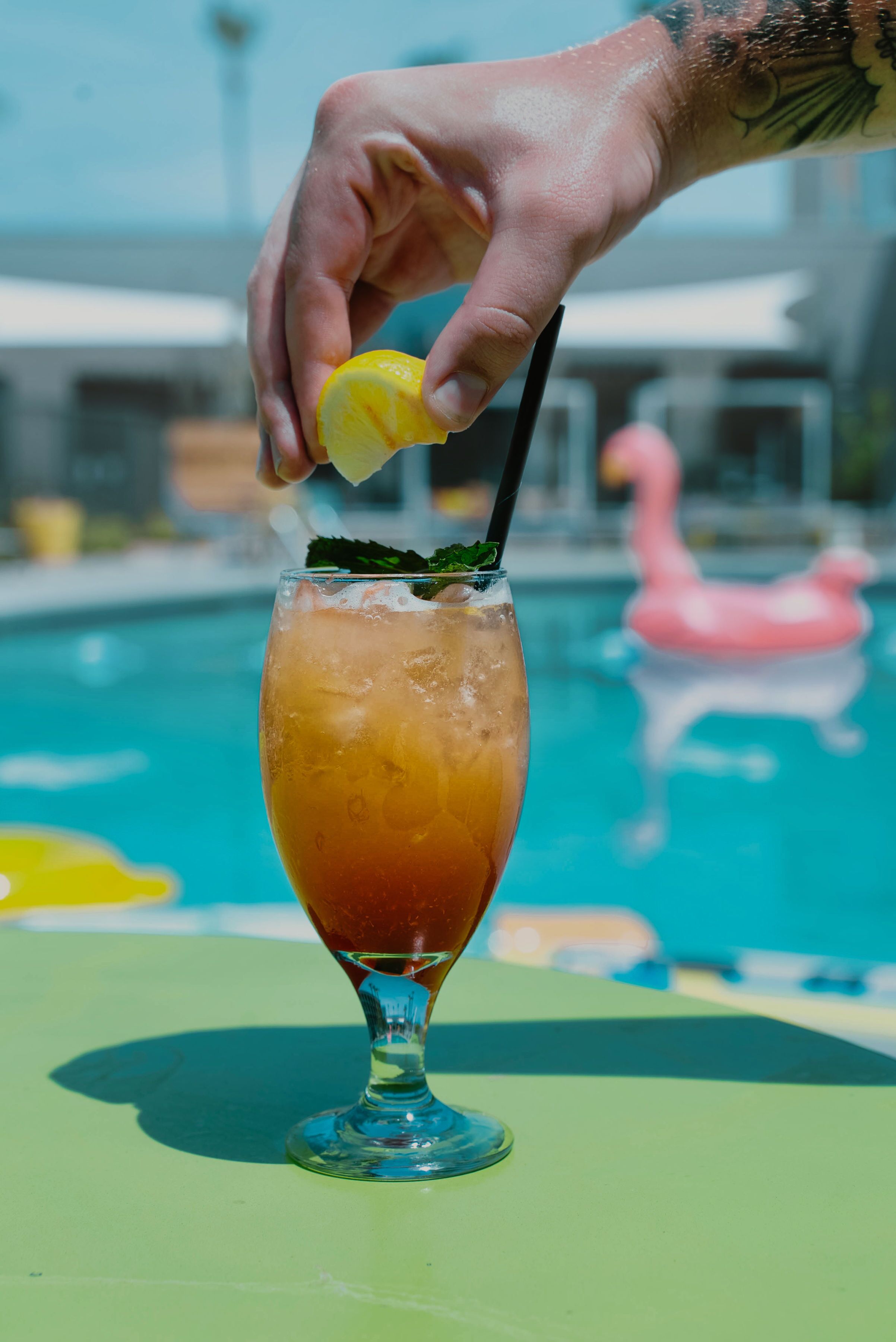 A cocktail in a tall glass with a lemon slice garnish, placed on a green table by a pool with a pink flamingo float.