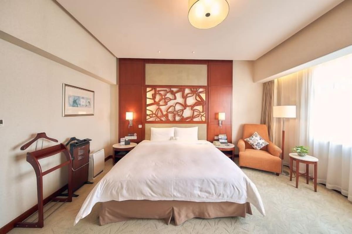 Habitación de hotel con una cama grande, dos mesitas de noche, una silla, una pequeña mesa, un radiador y un panel decorativo en la pared.