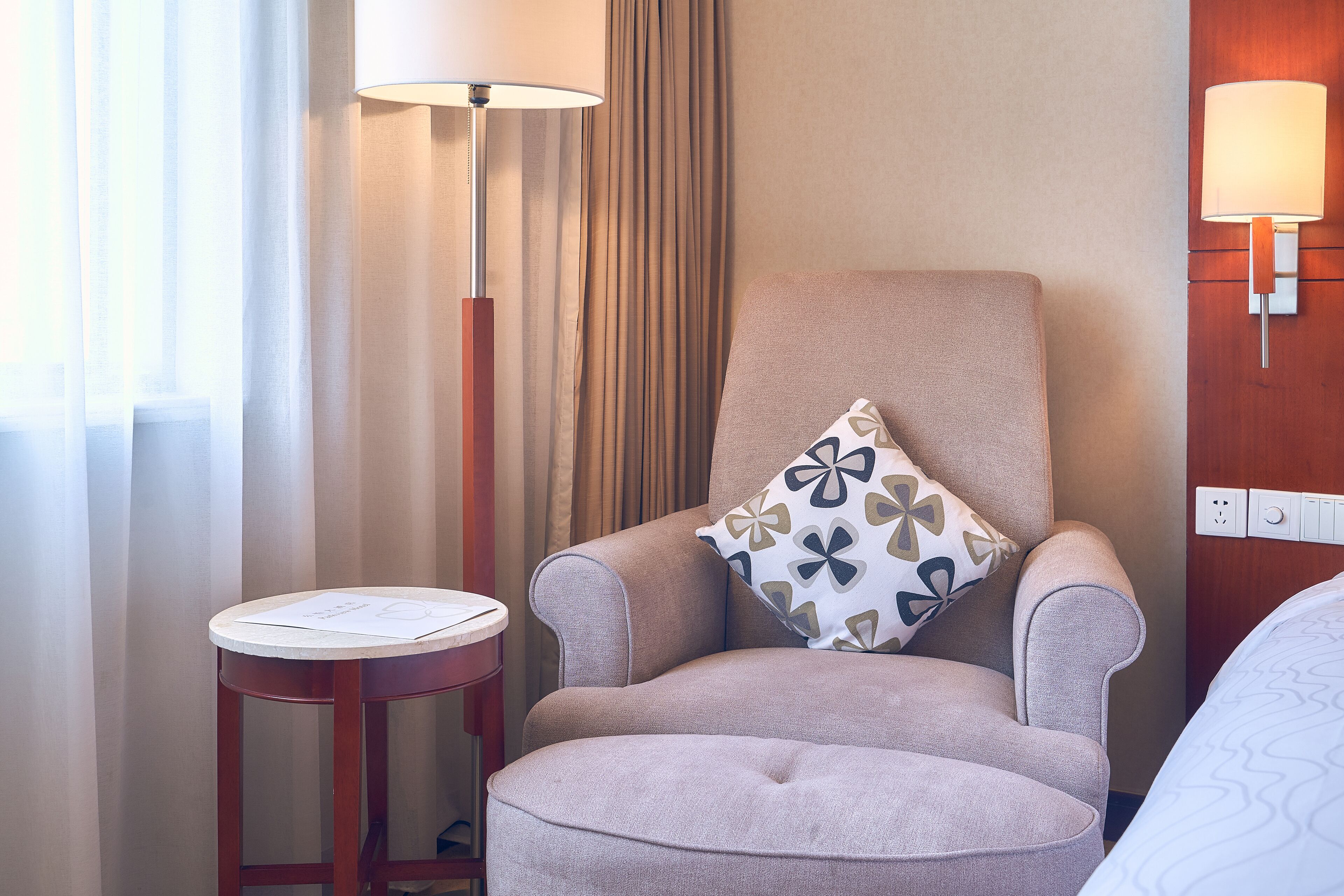A hotel room with a cushioned armchair, a small round table with a menu, a wall-mounted lamp, and a bed with a patterned bedspread.