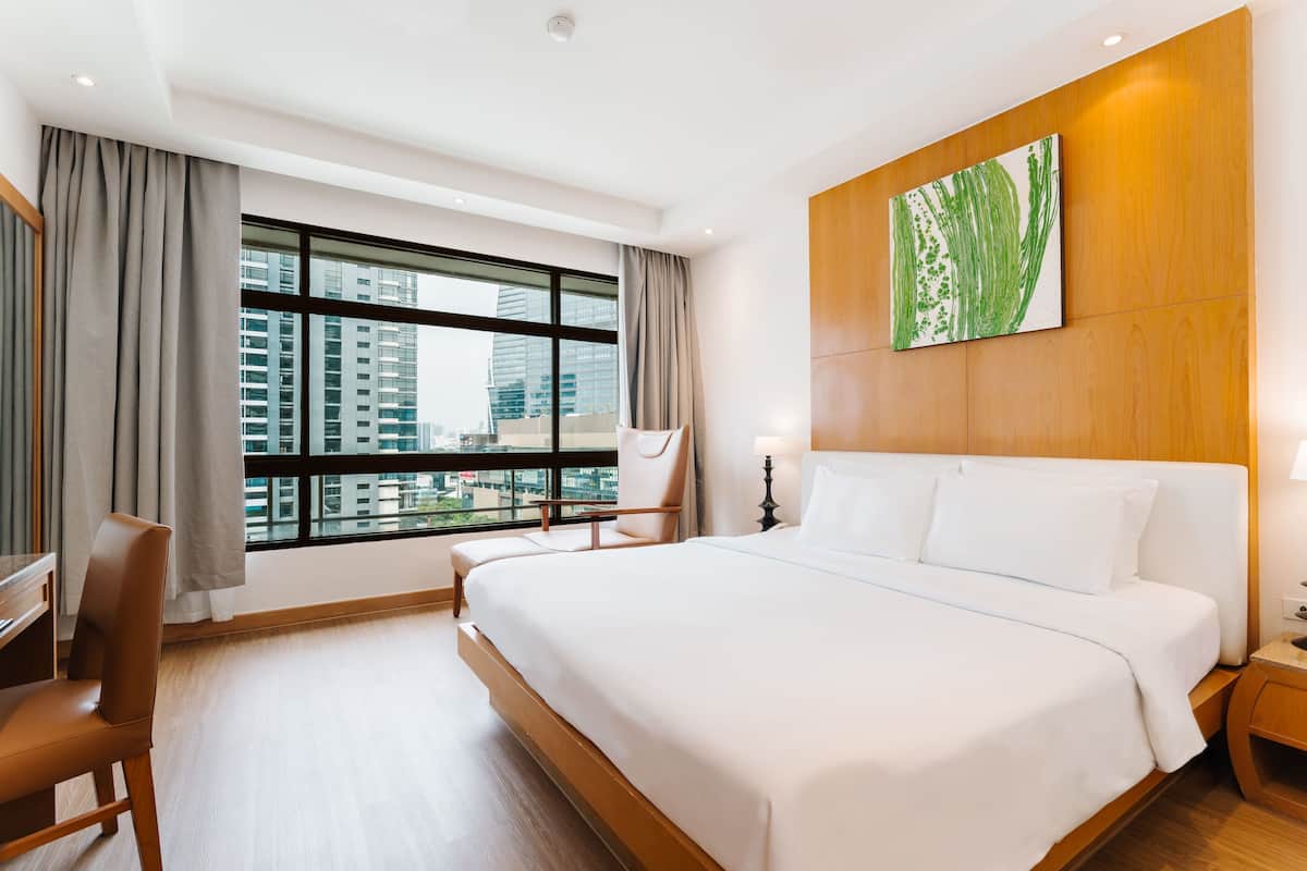 A hotel room with a large bed, a desk, a chair, a nightstand, and a window with a city view.