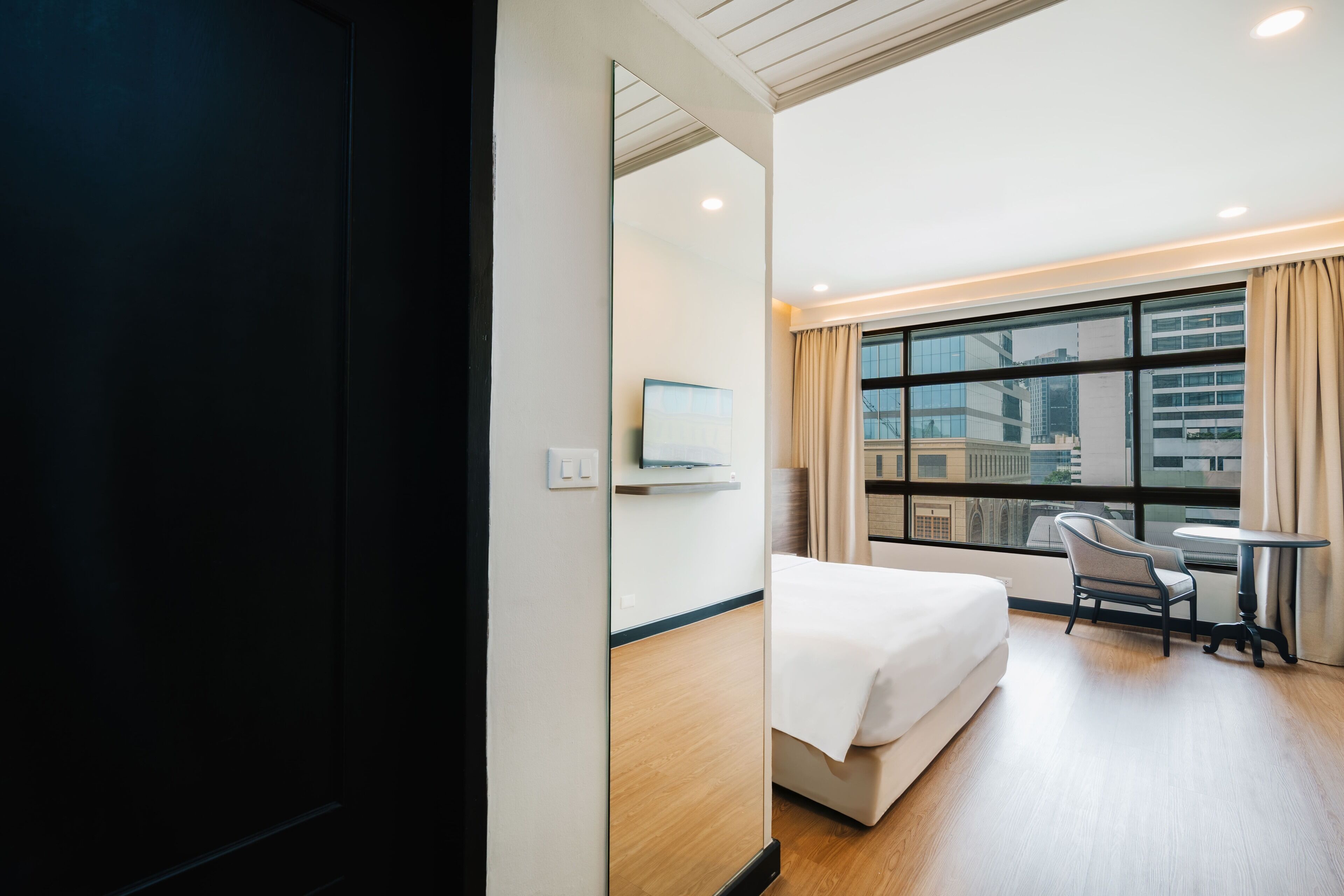 A hotel room with a bed, a chair, a small table, a TV, and a large window with curtains.